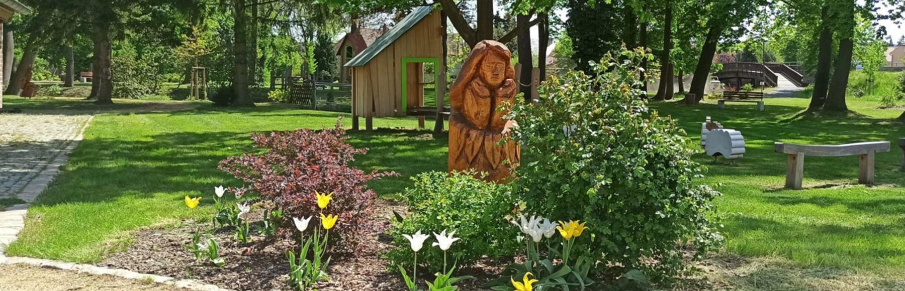 Blick auf den Spielplatz im Gutspark hinter dem Gemeindeamt Kodersdorf. Im Vordergrund sieht man eine aus Holz geschnitzte Figur einer Frau. Um die Figur herum blühen Tulpen.
