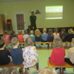 Zu sehen ist die Turnhalle unserer Kindertagesstätte. Auf mehreren Bänken verteilt sitzen die Kinder mit dem Rücken zu Kamera. Vor den Kindern steht ein Mann der Naturschutzstation. Er hält einen Vortrag zu Störchen anhand einer PowerPoint.